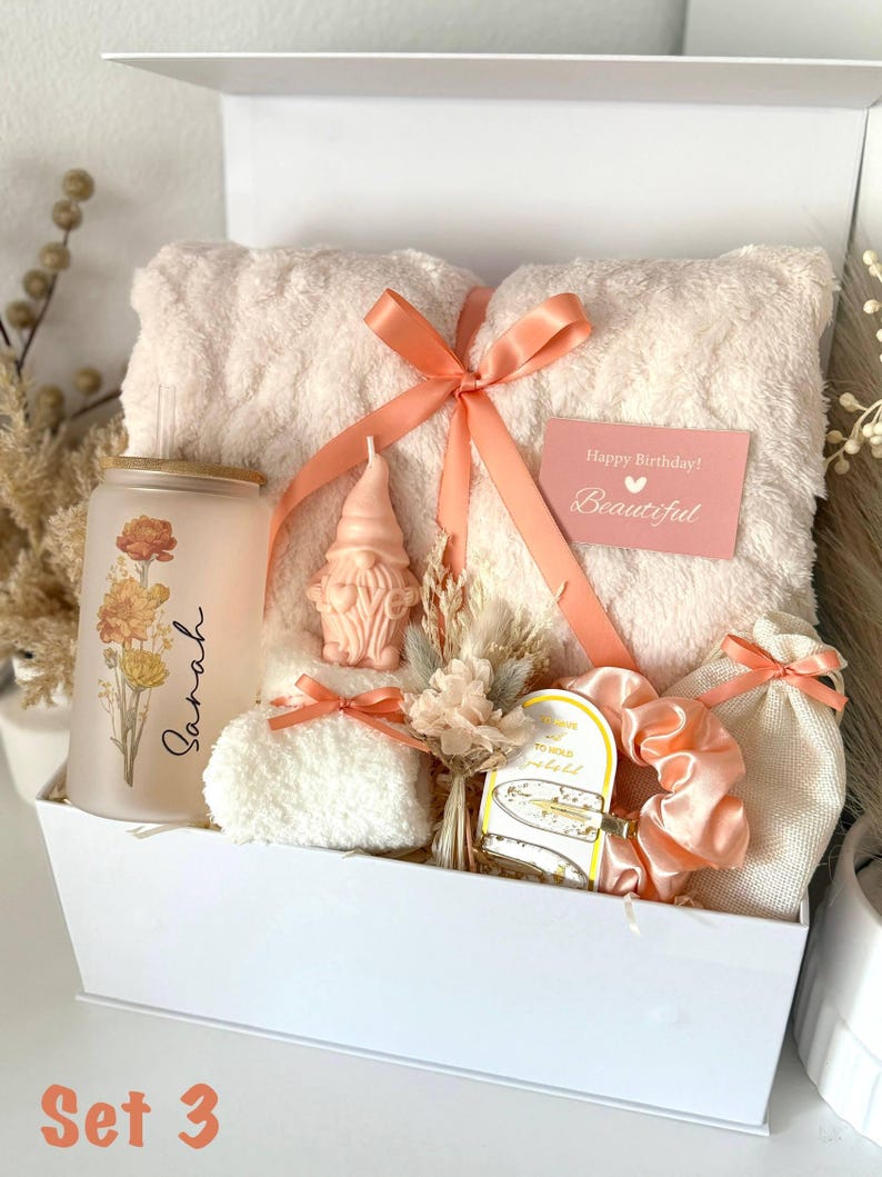 May include: A white gift box filled with a peach-colored towel, a glass tumbler with floral design and the name "Sarah", a gnome candle, scrunchies, and a small gift bag. A card reads "Happy Birthday! Beautiful".
