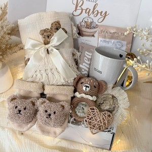 May include: A gift box labeled Hello Baby containing a cream-colored muslin blanket with bear embroidery and a satin ribbon, beige bear-faced socks, a gray mug labeled mom, a crochet bear rattle, a woven heart, and a gold-toned spoon.