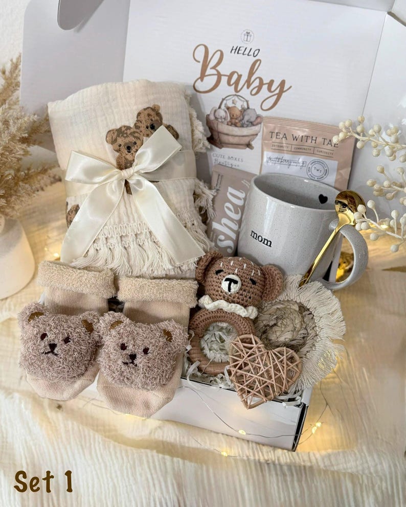 May include: A gift box labeled Hello Baby containing a cream-colored muslin blanket with bear embroidery and a satin ribbon, beige bear-faced socks, a gray mug labeled mom, a crochet bear rattle, a woven heart, and a gold-toned spoon.