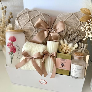 May include: A white gift box containing a beige textured pillow, a glass with a floral design and the name "Sarah", a white towel with a bow, a candle, soap, bath salts, and decorative elements. The set is tied with a brown satin ribbon.