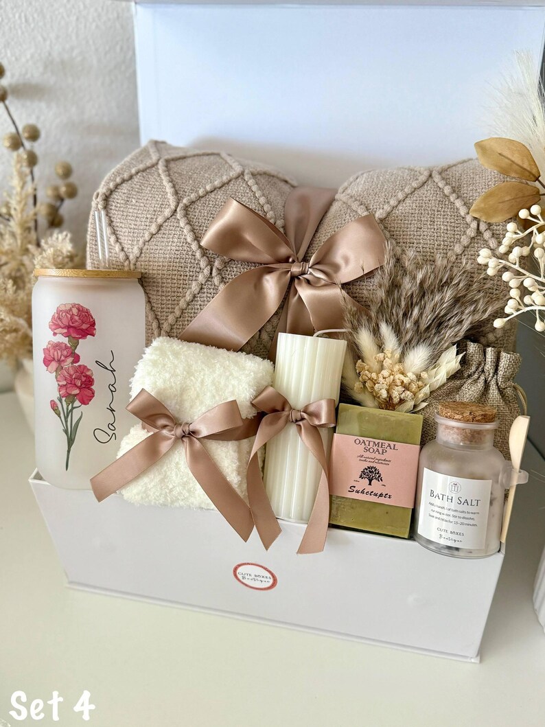 May include: A white gift box containing a beige textured pillow, a glass with a floral design and the name "Sarah", a white towel with a bow, a candle, soap, bath salts, and decorative elements. The set is tied with a brown satin ribbon.