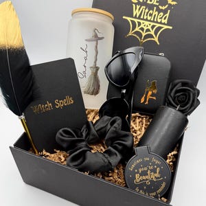 May include: A black gift box containing a frosted glass tumbler with a witch hat and broom graphic, a black notebook labeled Witch Spells, a black feather pen, black heart-shaped sunglasses, a black scrunchie, a black candle, and a round mirror.