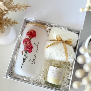 May include: A white gift box contains a frosted glass tumbler with a floral design and the name "Sarah", a small candle, and a white fluffy towel tied with a gold ribbon. The candle has a label that reads "A Moment of Peace and Comfort".