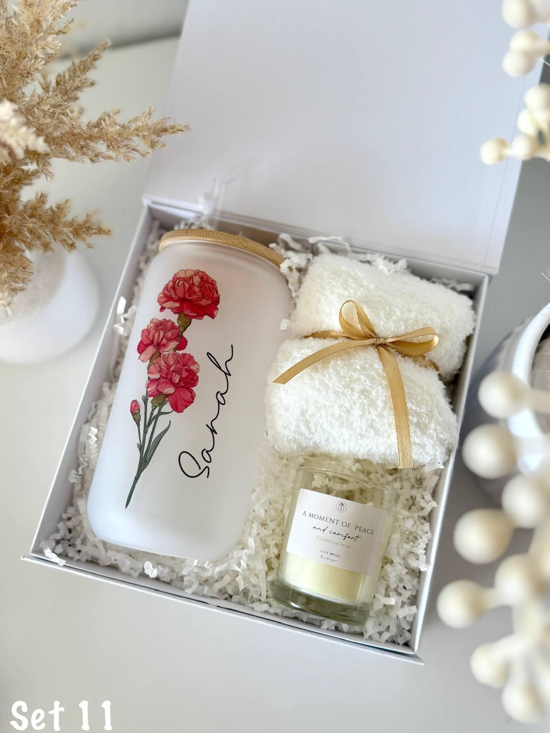 May include: A white gift box contains a frosted glass tumbler with a floral design and the name "Sarah", a small candle, and a white fluffy towel tied with a gold ribbon. The candle has a label that reads "A Moment of Peace and Comfort".