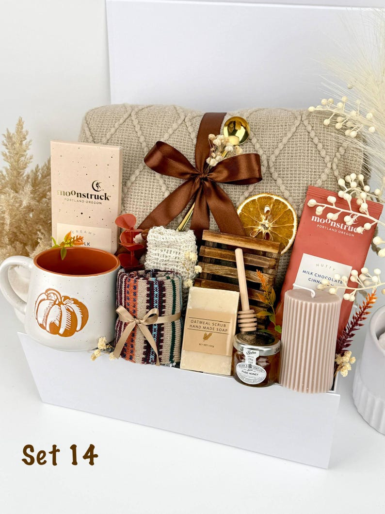 May include: A gift set containing a beige knit blanket with a brown ribbon, a ceramic mug with a pumpkin graphic, two Moonstruck chocolate bars, a bar of oatmeal scrub soap, a wooden honey dipper, a jar of honey, and a ribbed pillar candle.