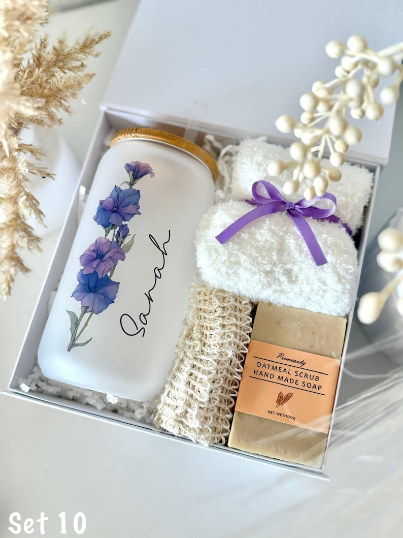 May include: A white gift box containing a glass tumbler with a floral design and the name "Sarah", a white washcloth with a purple ribbon, a loofah, and a bar of oatmeal scrub soap. The set is labeled "Set 10".