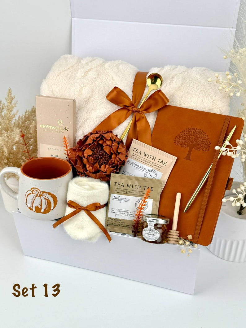 May include: A gift set containing a white textured throw blanket, a rust-colored notebook with a tree emblem, a ceramic mug with a pumpkin graphic, a gold spoon, a gold pen, two tea sachets labeled Tea With Tae, a jar of honey, and a honey dipper.