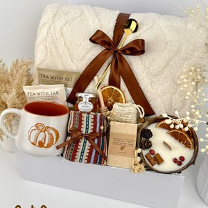 May include: A gift set containing a cream-colored textured throw blanket tied with a brown ribbon, a gold spoon, a pumpkin-print mug, tea packets, a jar of honey, a patterned cloth, oatmeal soap, and a candle in a coconut shell with dried fruit.