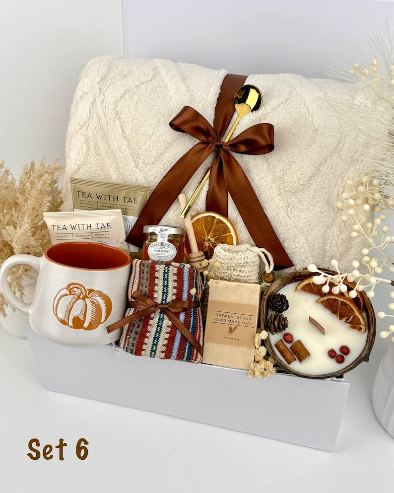 May include: A gift set containing a cream-colored textured throw blanket tied with a brown ribbon, a gold spoon, a pumpkin-print mug, tea packets, a jar of honey, a patterned cloth, oatmeal soap, and a candle in a coconut shell with dried fruit.