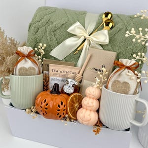 May include: A gift set containing a sage green textured throw blanket with a cream ribbon, two ribbed mugs, two burlap pouches with heart designs, a ceramic orange pumpkin, a stacked pumpkin candle, a honey dipper, tea packets, and chocolate.