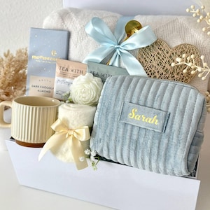 May include: A gift set containing a blue ribbed velvet pouch with the name Sarah embroidered in gold, a cream ribbed mug, a white knit blanket, a Moonstruck dark chocolate bar, two tea packets, a white flower, and a gold heart ornament.