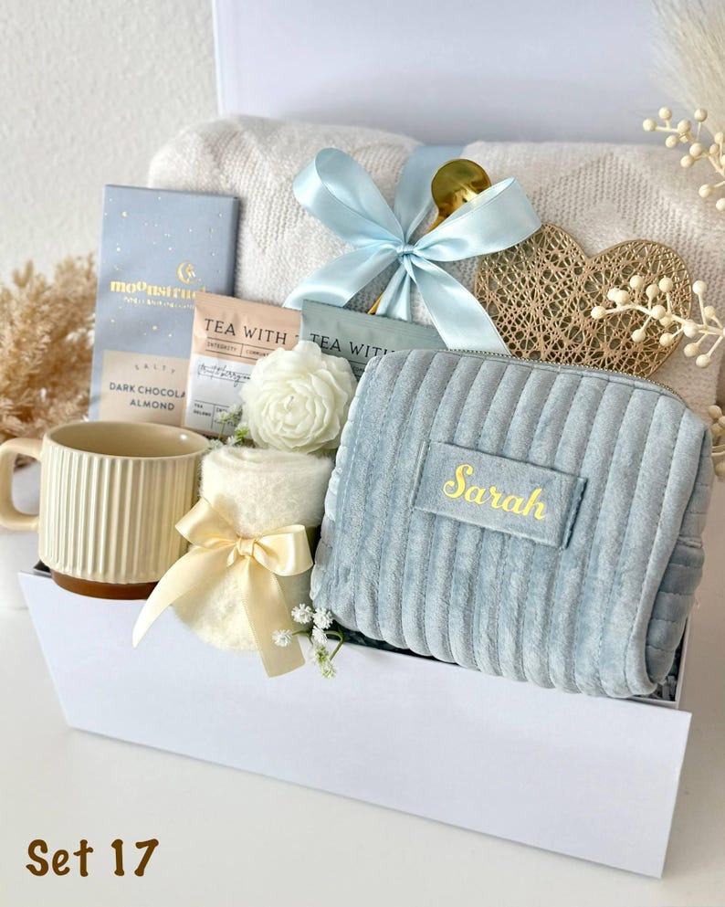 May include: A gift set containing a blue ribbed velvet pouch with the name Sarah embroidered in gold, a cream ribbed mug, a white knit blanket, a Moonstruck dark chocolate bar, two tea packets, a white flower, and a gold heart ornament.