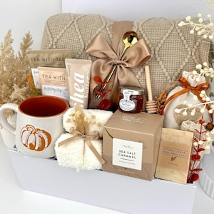May include: A gift set containing a beige cable-knit blanket with a satin bow, a ceramic mug with a pumpkin graphic, tea packets, shea butter, a jar of honey with a wooden dipper, sea salt caramel cookies, oatmeal soap, and a small pouch.