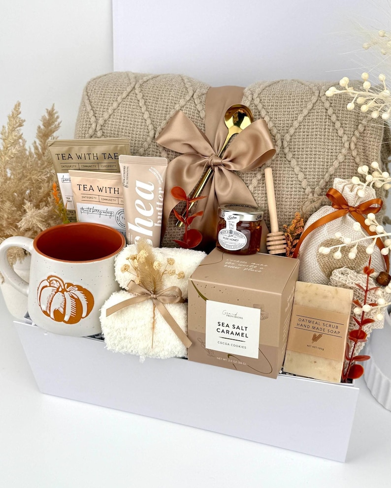 May include: A gift set containing a beige cable-knit blanket with a satin bow, a ceramic mug with a pumpkin graphic, tea packets, shea butter, a jar of honey with a wooden dipper, sea salt caramel cookies, oatmeal soap, and a small pouch.