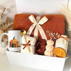 May include: A white gift box containing a folded burnt orange knit blanket tied with a cream ribbon, a white mug with a pumpkin illustration, a white fuzzy cloth with a gold spoon, a brown pinecone, a stacked pumpkin candle, and a dried orange slice.