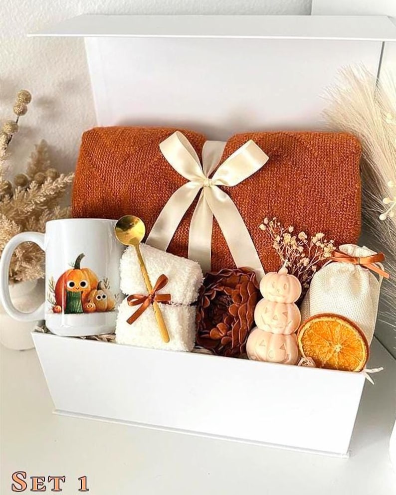 May include: A white gift box containing a folded burnt orange knit blanket tied with a cream ribbon, a white mug with a pumpkin illustration, a white fuzzy cloth with a gold spoon, a brown pinecone, a stacked pumpkin candle, and a dried orange slice.