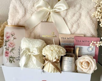 Op de afbeelding: Een witte geschenkdoos gevuld met zelfzorgartikelen. Inclusief een zachte deken, een met bloemen versierd glas, een kleine handdoek met een strik, een kaars, badzout, thee en een roosvormige zeep. De doos is vastgebonden met een grote witte strik.
