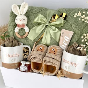 May include: A gift set containing a sage green textured blanket tied with a ribbon, a crochet bunny rattle with the name Alex, two mugs labeled daddy and mommy with names George and Maria, beige baby shoes with bear faces, and a tube of shea cream.