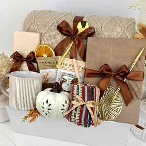 May include: A gift set containing a beige cable-knit blanket with a brown ribbon, a ribbed ceramic mug, a white pumpkin-shaped candle, tea packets, a small honey jar, a notebook with a gold pen, and patterned socks. The text Set 17 is visible.