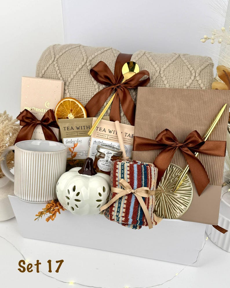 May include: A gift set containing a beige cable-knit blanket with a brown ribbon, a ribbed ceramic mug, a white pumpkin-shaped candle, tea packets, a small honey jar, a notebook with a gold pen, and patterned socks. The text Set 17 is visible.