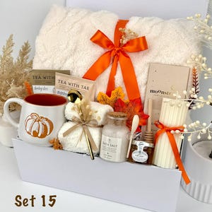 May include: A gift set in a white box containing a cream-colored textured blanket tied with an orange ribbon, a mug with a pumpkin graphic, two tea packets, a gold spoon, bath salts, a jar of honey with a dipper, and a ribbed pillar candle.