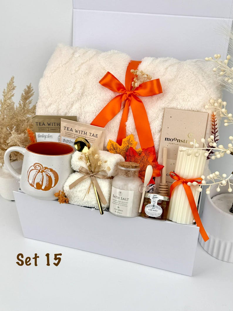 May include: A gift set in a white box containing a cream-colored textured blanket tied with an orange ribbon, a mug with a pumpkin graphic, two tea packets, a gold spoon, bath salts, a jar of honey with a dipper, and a ribbed pillar candle.