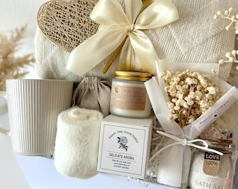 May include: A gift basket filled with self-care items. It includes a white mug, a rolled-up blanket, a candle, bath salts, a small bouquet of dried flowers, and a decorative heart. The items are arranged in a white box and tied with a cream-colored ribbon.