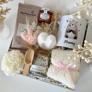 K&ouml;nnte beinhalten: Geschenkbox mit einer Tasse mit einer B&auml;renillustration und dem Wort "Mommy", einem Honigglas, einer Schokoladenpackung, Badesalz, einer Kerze, einer herzf&ouml;rmigen Badekugel, einem Honigl&ouml;ffel aus Holz und flauschigen Socken mit rosa Schleife. Die Box ist mit "Set 6" beschriftet.
