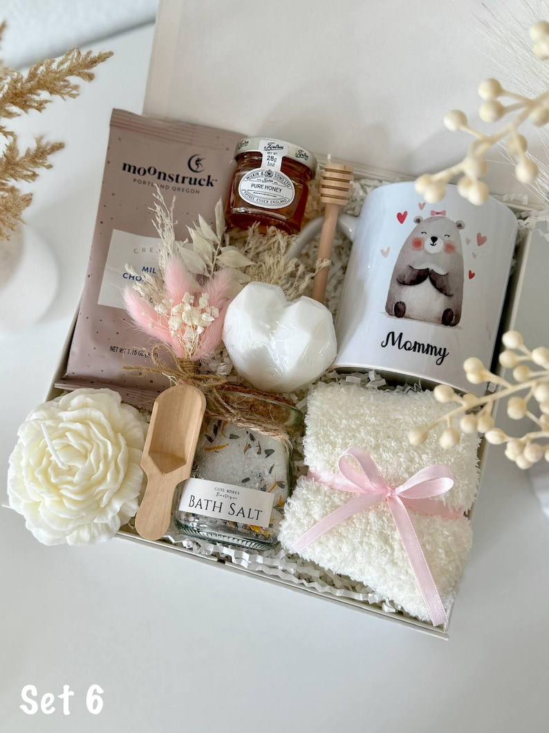 K&ouml;nnte beinhalten: Geschenkbox mit einer Tasse mit einer B&auml;renillustration und dem Wort "Mommy", einem Honigglas, einer Schokoladenpackung, Badesalz, einer Kerze, einer herzf&ouml;rmigen Badekugel, einem Honigl&ouml;ffel aus Holz und flauschigen Socken mit rosa Schleife. Die Box ist mit "Set 6" beschriftet.