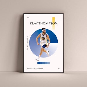May include: A basketball player wearing a blue and gold jersey with the number 11 on it. The player is dribbling the ball and is in mid-air. The background is a blue and white circle. The text "KLAY THOMPSON" is at the top of the image. The text "2011/ROUND 1/PICK 11" is on the left side of the image. The text "GOLDEN STATE WARRIORS" is at the bottom of the image. The text "SG" is on the right side of the image.