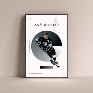 Può includere: Un poster in bianco e nero con un giocatore di hockey che indossa una maglia dei Los Angeles Kings. Il poster presenta il nome del giocatore, Anže Kopitar, e le sue informazioni di draft: 2005 Round 1 Pick 11. Il poster include anche il logo della squadra e la posizione del giocatore, C.
