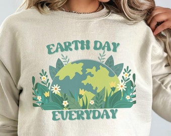 Puede incluir: Una sudadera de color beige claro con un gráfico verde y azul de la Tierra rodeado de flores y hojas. El texto "Earth Day Everyday" está impreso en letras verdes en la sudadera.