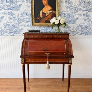 May include: A mahogany roll-top desk featuring a green marble top, three small upper drawers, and a central pull-out writing surface. The desk is accented with gold-toned metal hardware, including a key with a cream-colored tassel.