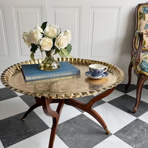 May include: A round brass tray table with a scalloped edge and intricate etched patterns, supported by dark wooden legs with brass feet. On the tray sits a glass vase of white roses, a blue book titled The Grammar of Chinese Ornament, and a blue and white teacup.
