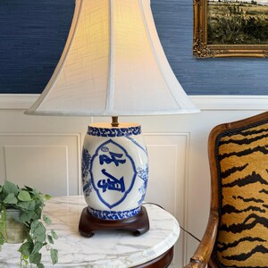 Puede incluir: Una lámpara de mesa con una pantalla de tela blanca en forma de campana y una base de cerámica con motivos florales azules y blancos y caracteres de caligrafía china. La lámpara se asienta sobre una base de madera oscura con un remate de latón.