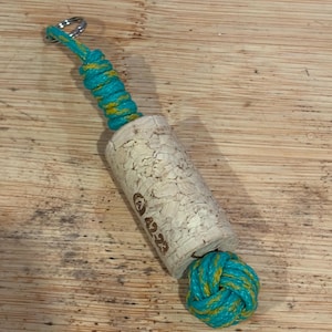 Puede incluir: Un tapón de botella de vino hecho de corcho con un diseño de cuerda turquesa y amarilla. El corcho tiene los números "42.23" impresos. La cuerda está anudada en la parte inferior y tiene un anillo de metal en la parte superior.