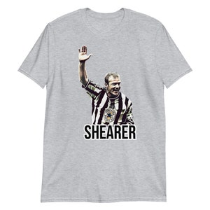 May include: A gray t-shirt with a black and white image of a soccer player raising his hand in the air. The player is wearing a black and white striped jersey with a star on the chest. The text "SHEARER" is printed in black below the image.