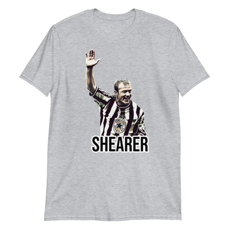 May include: A gray t-shirt with a black and white image of a soccer player raising his hand in the air. The player is wearing a black and white striped jersey with a star on the chest. The text "SHEARER" is printed in black below the image.