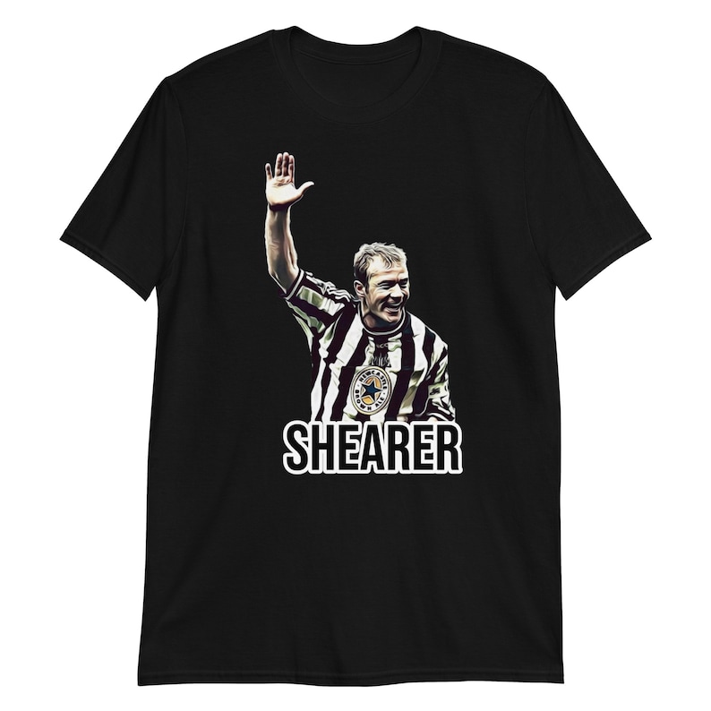 May include: Black t-shirt with a white graphic of a soccer player in a black and white striped jersey raising his hand in the air. The text "SHEARER" is printed below the image.