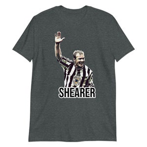 May include: A dark heather grey t-shirt with a graphic of Alan Shearer, a famous English footballer, in a black and white striped jersey. The text "SHEARER" is printed below the graphic.