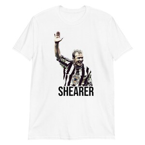 May include: White t-shirt with a black and white image of a soccer player raising his hand in the air. The player is wearing a black and white striped jersey with a team logo. The text "SHEARER" is printed below the image.