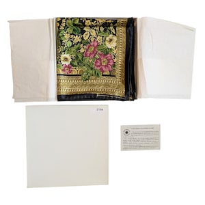 May include: A square scarf with a black background featuring a floral pattern of pink, white, and yellow flowers with green leaves. The scarf has a gold-toned decorative border and is presented in an open white box with an informational card.