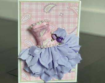 May include: A handmade greeting card with a 3D paper dress design. The dress is pink with a white and purple floral pattern and a lavender ruffle skirt. The card has a pink and white gingham background with a paisley pattern.