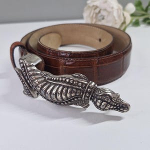 May include: Brown leather belt with a silver-toned crocodile-shaped buckle. The belt has a textured, embossed pattern. The buckle is detailed with scales and teeth. The belt is rolled up on a white surface.
