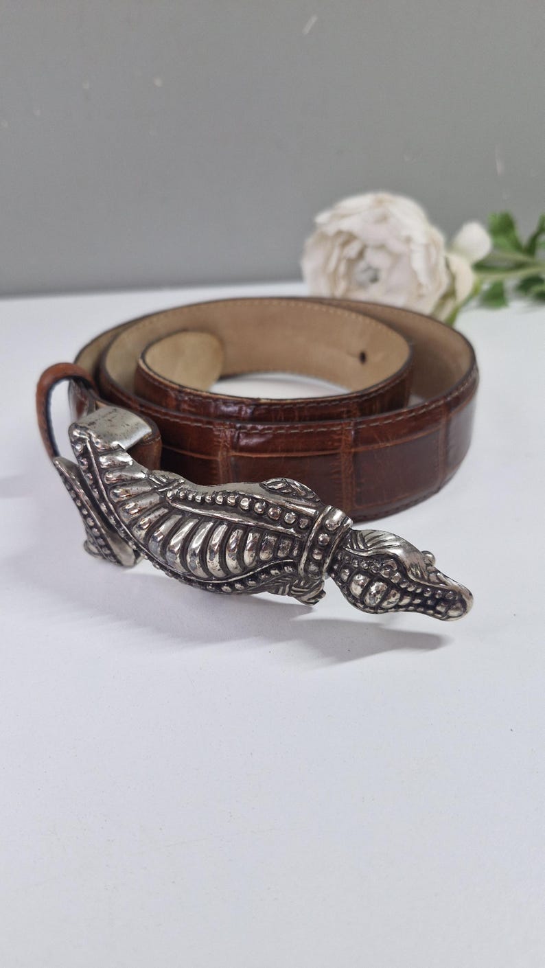 May include: Brown leather belt with a silver-toned crocodile-shaped buckle. The belt has a textured, embossed pattern. The buckle is detailed with scales and teeth. The belt is rolled up on a white surface.