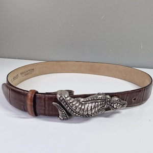 May include: A brown leather belt with a silver-colored alligator-shaped buckle. The belt has a textured, crocodile-like pattern and a light brown interior. The buckle is detailed with scales and teeth.