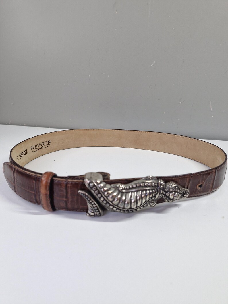 May include: A brown leather belt with a silver-colored alligator-shaped buckle. The belt has a textured, crocodile-like pattern and a light brown interior. The buckle is detailed with scales and teeth.