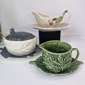 Könnte beinhalten: Drei Keramik-Saucieren mit Blattmotiven. Eine ist weiß mit rosa Blüten, eine ist grün und eine ist weiß mit einem grauen Deckel.