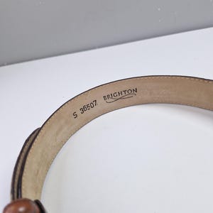May include: A brown leather belt with a woven design and a small, round buckle. The brand name "BRIGHTON" and the number "S 36507" are visible on the inside of the belt.