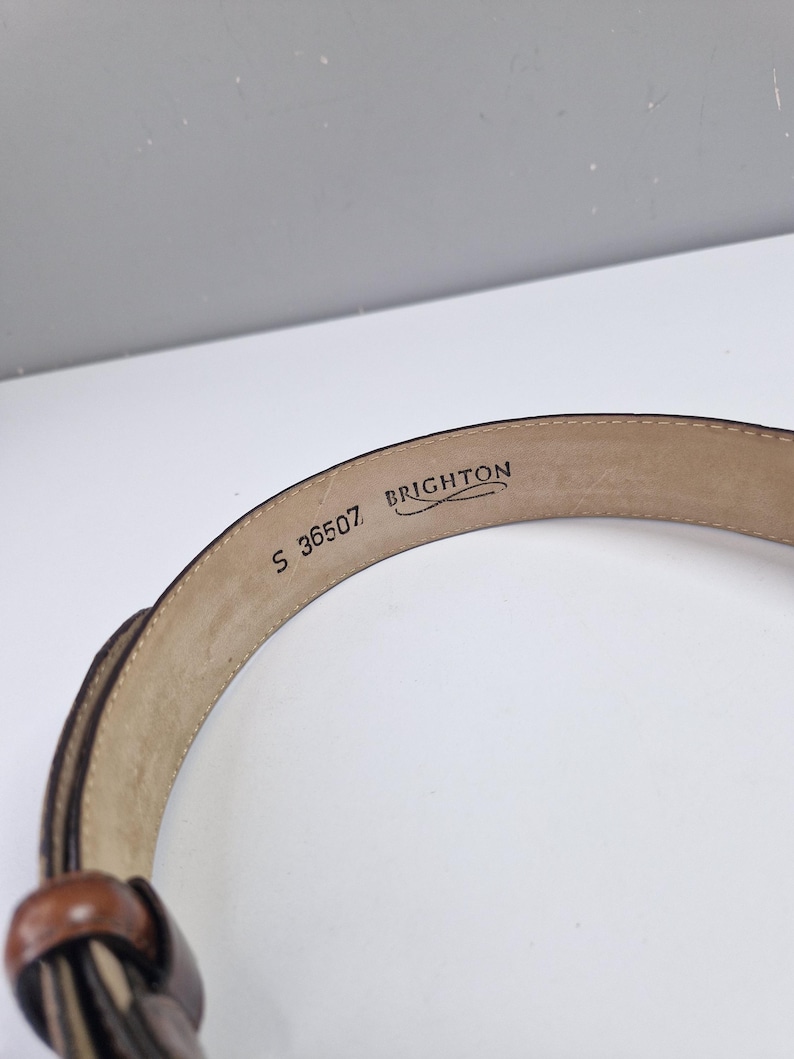 May include: A brown leather belt with a woven design and a small, round buckle. The brand name "BRIGHTON" and the number "S 36507" are visible on the inside of the belt.